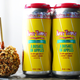 A caramel apple on a table beside two bright yellow cans of "A Bushel of Apples" beer